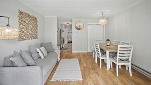 Una sala de estar moderna con un sofá gris, cojines blancos, una mesa de comedor de madera con sillas blancas y una alfombra blanca.