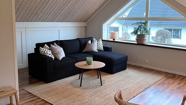 A loft-style room with a sloped ceiling, a black sectional sofa, a round wooden coffee table, a wicker chair, and a large window with a view of a house. - Cozy apartment in own house with terrace near Lassalyckan (Västra Götalands län)