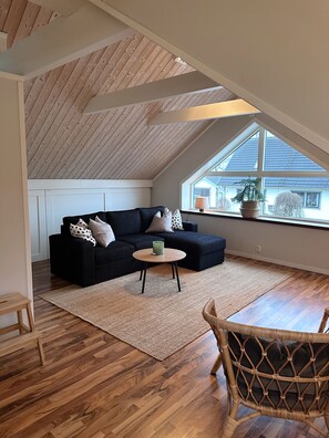 Een loft-achtige kamer met een schuine plafond, een zwarte hoekbank, een ronde houten salontafel, een rieten stoel en een groot raam met uitzicht op een huis.