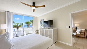 A hotel room with a large bed, a television on a cabinet, a ceiling fan, and a view of palm trees through a sliding glass door. - Beach Club 210 - King/King/Queen/Sofa Sleeper - 2nd FL Corner by the Club- 8 max (Gulf Shores)
