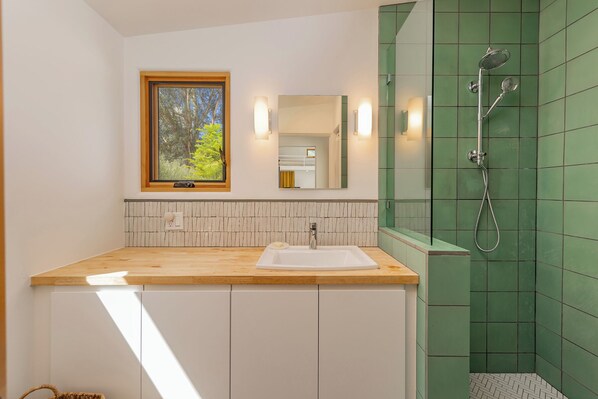 A bathroom with a wooden vanity, a white sink, a mirror, and a green-tiled shower area.
