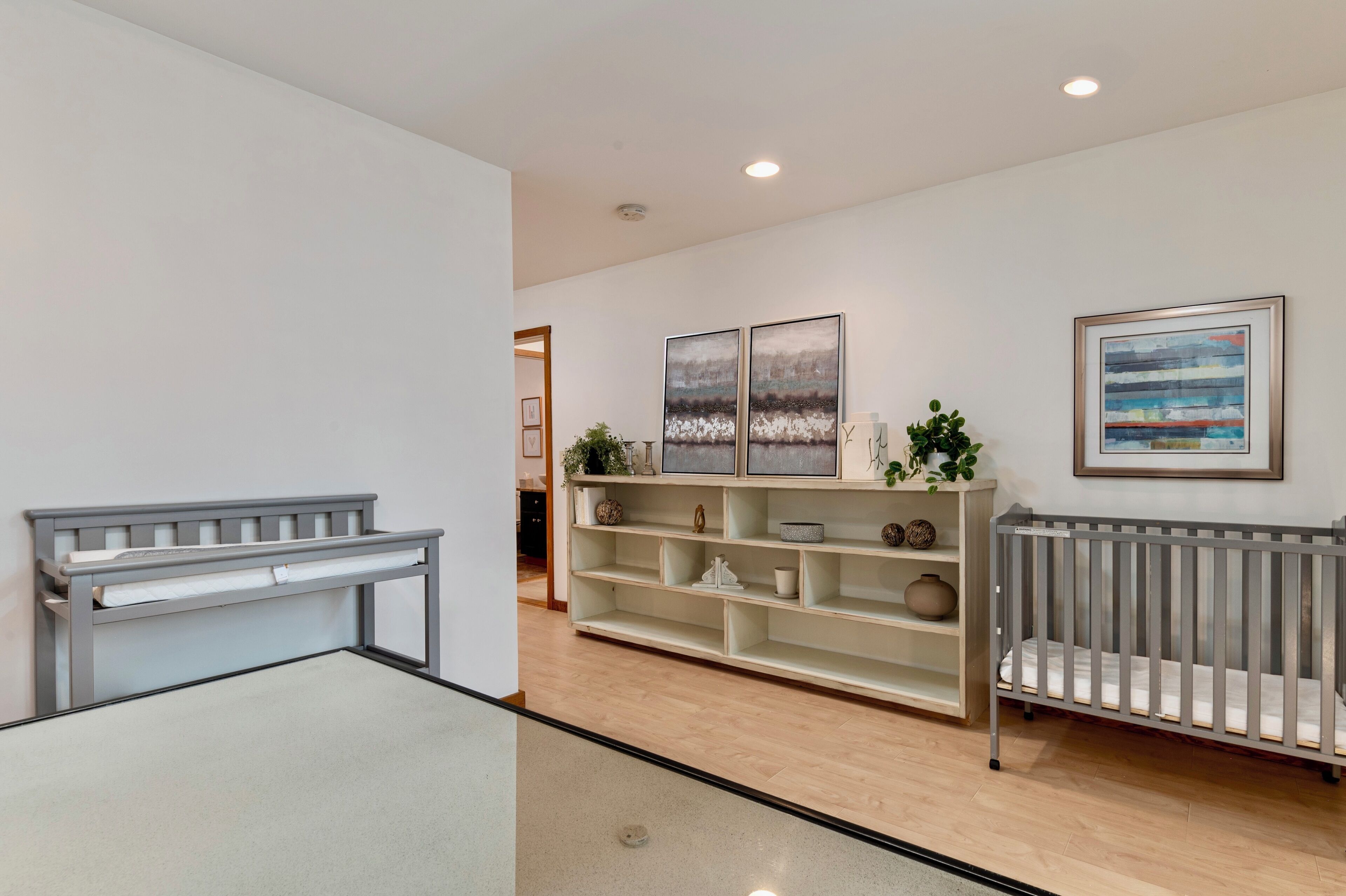 A modern interior with a grey crib, a white shelving unit with decorative items, and a framed abstract painting on the wall.