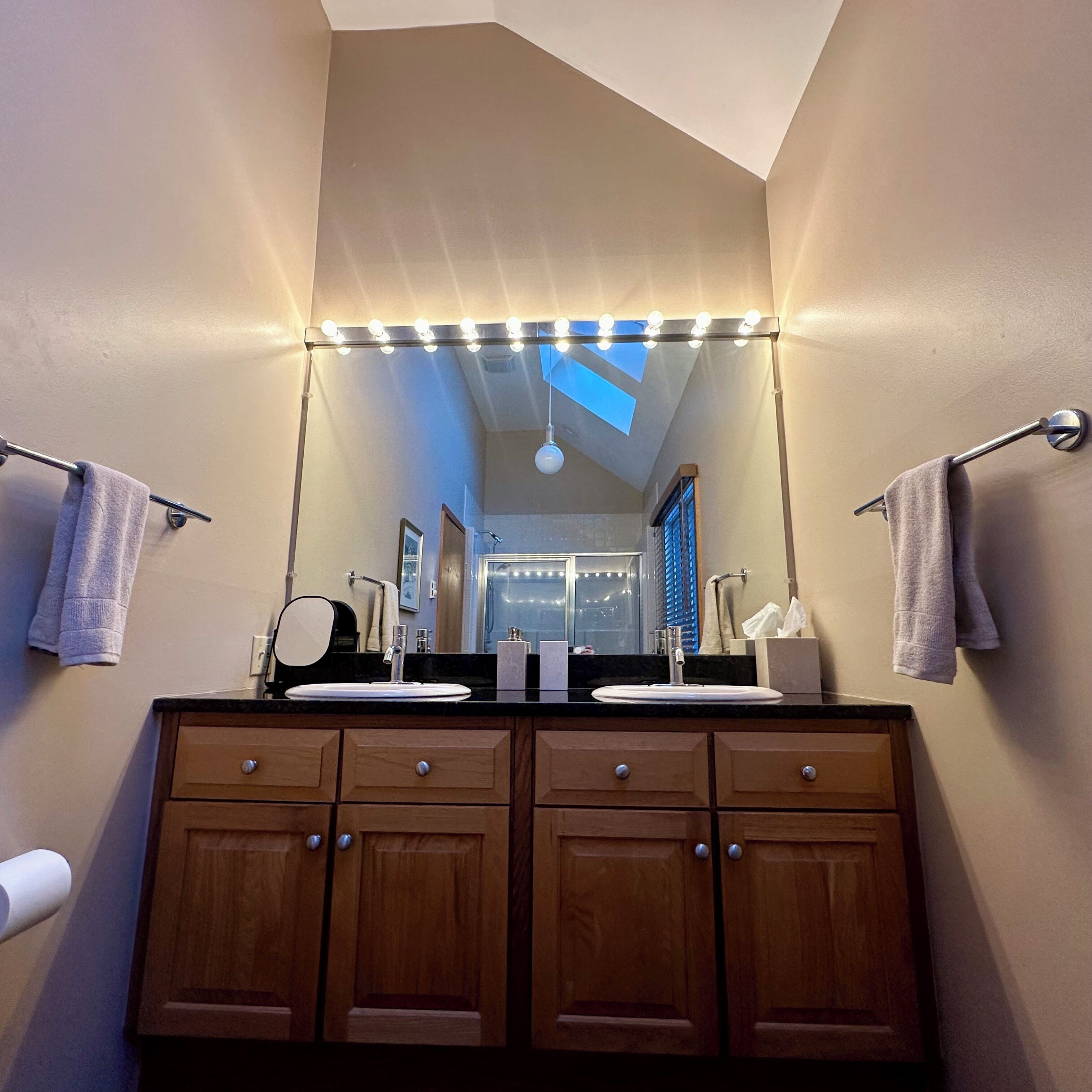 A bathroom with two sinks, a mirror with lights, wooden cabinets, and towels on towel bars.