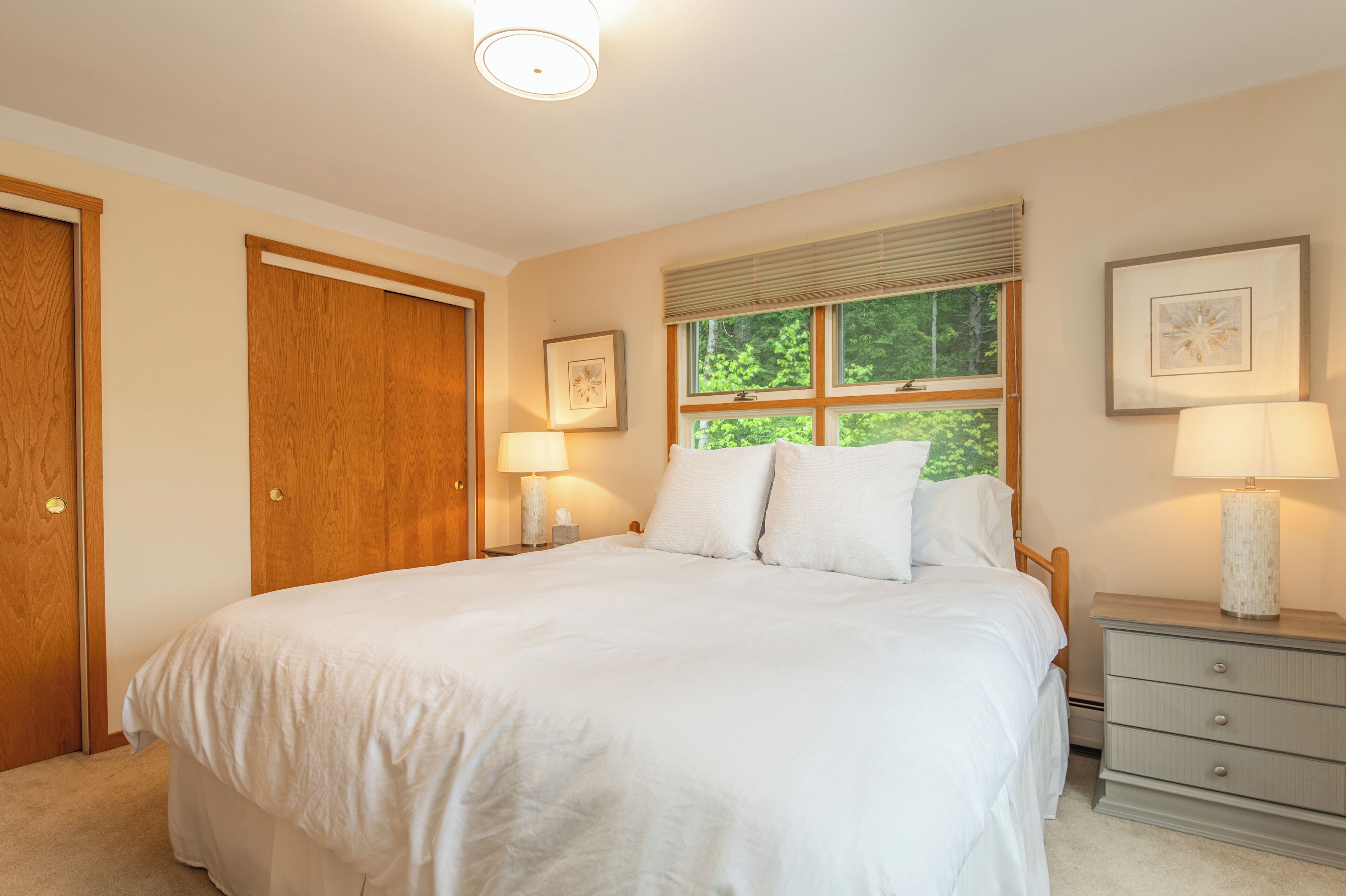 A bedroom with a large bed, two bedside tables with lamps, a window with blinds, and a door.