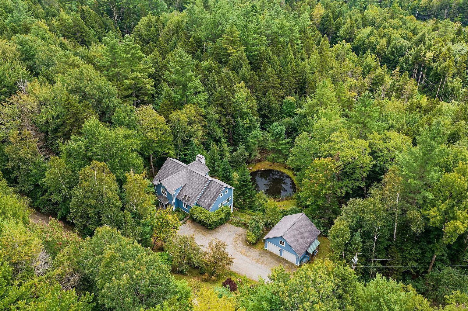Une vue aérienne d’une maison entourée d’une forêt dense, avec un étang visible et une allée bien dégagée menant à l’entrée principale.