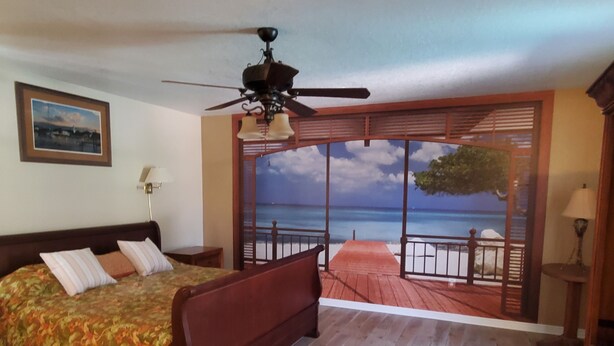 A bedroom with a wooden bed, a ceiling fan, a large window with a view of the sea, and a wooden floor. - Butterfly House, close to beaches, restaurants, live entertainment & shopping (Sarasota)