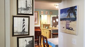A dining area with wooden chairs and a table, framed lighthouse paintings on the wall, and a kitchen in the background. - Butterfly House, close to beaches, restaurants, live entertainment & shopping (Sarasota)