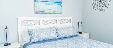 A bedroom with a white headboard, a bed with blue patterned bedding, a nightstand with a lamp, and a wall art of a beach scene.