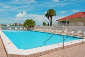 A rectangular swimming pool with a white border and a brick walkway. There are lounge chairs along the poolside, and a palm tree is visible in the background.