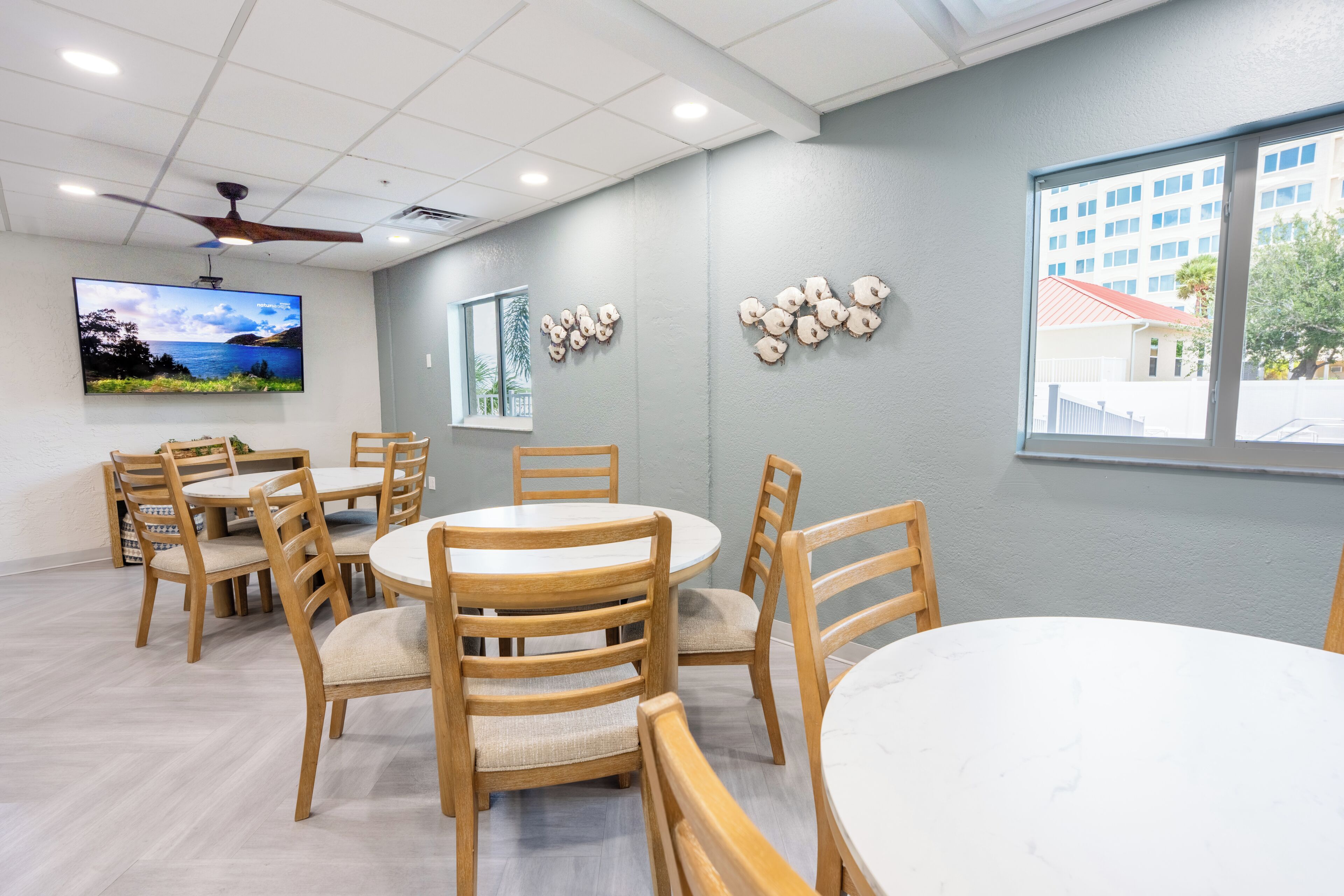 A conference room with round tables and wooden chairs, a flat-screen TV on the wall, and a window with a view of buildings and trees.