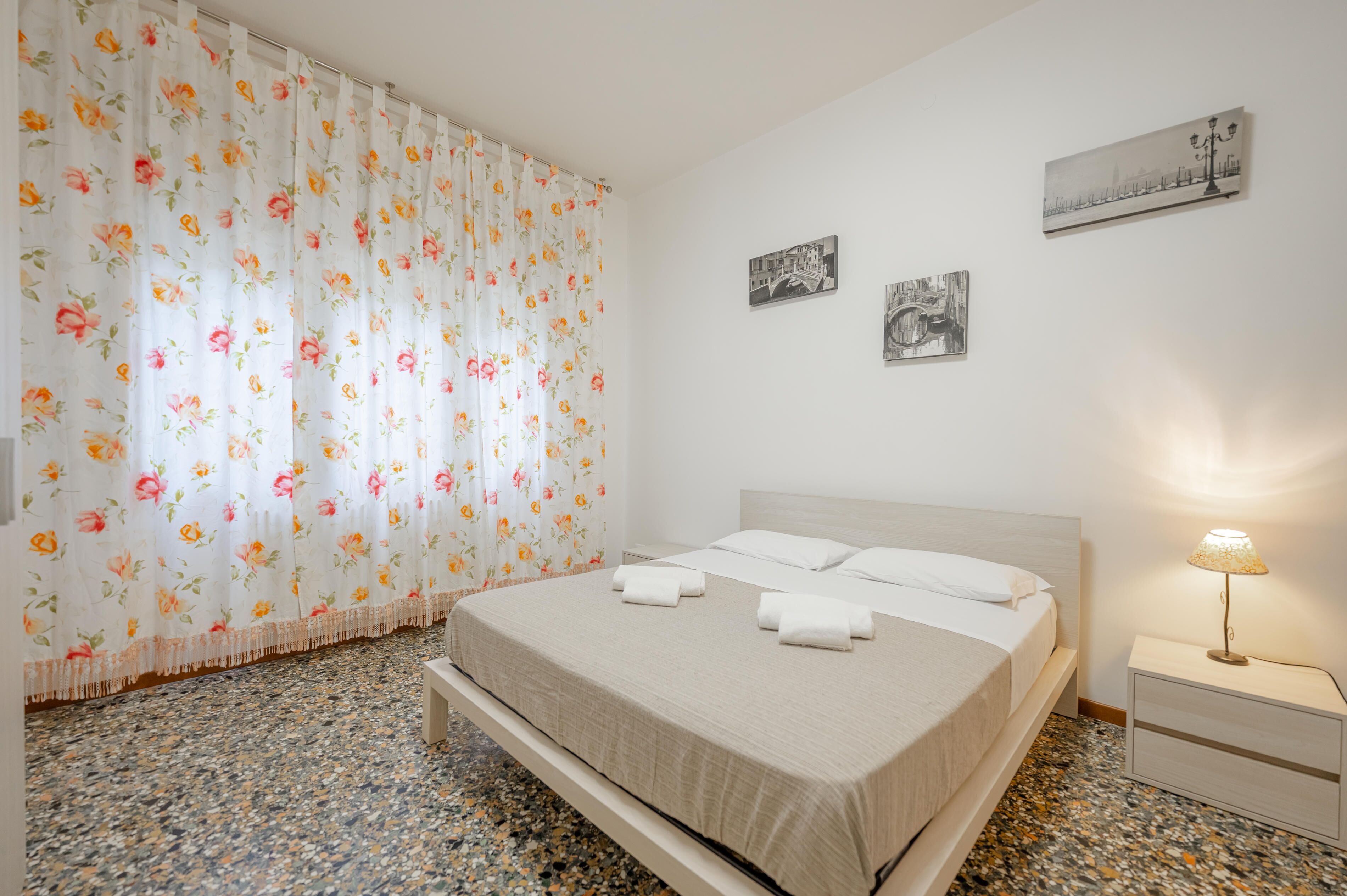 A neatly made bed with white linens and a beige blanket, a nightstand with a lamp, and a floral curtain.