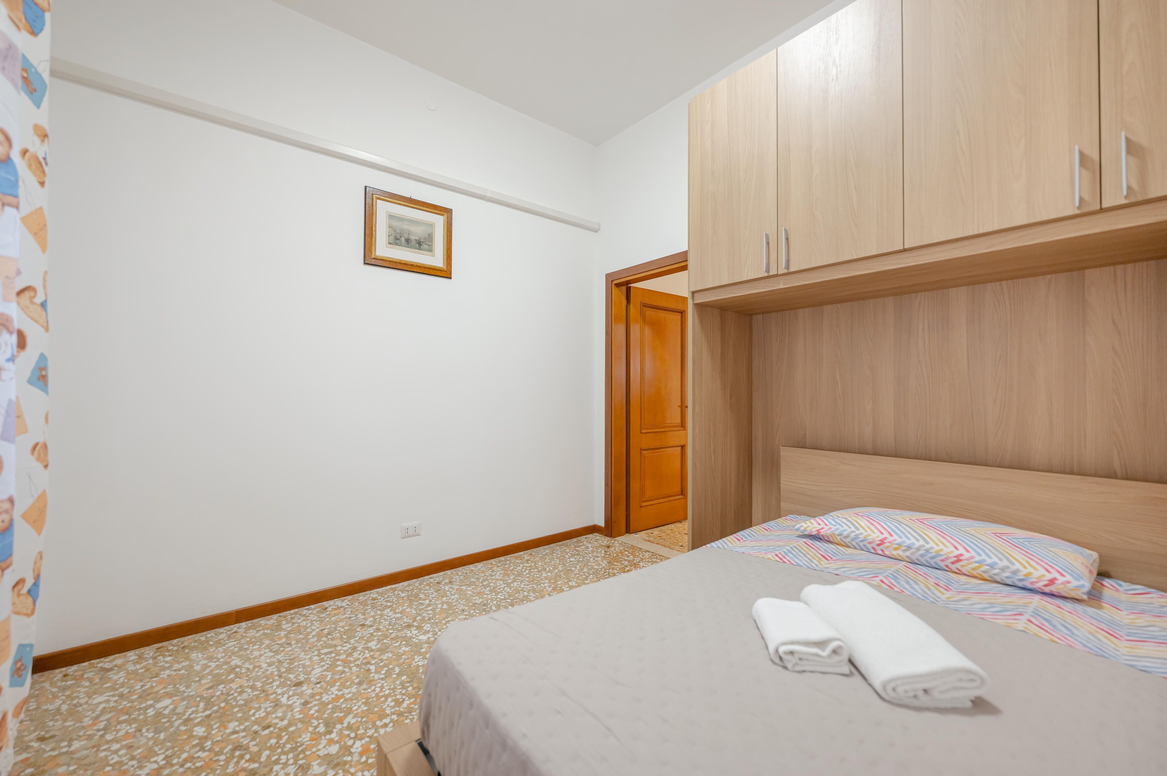 Un dormitorio con una cama, un armario de madera, un cuadro enmarcado en la pared y una puerta que da a otra habitación.
