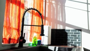 A kitchen sink with a modern faucet, a window with orange curtains, and a brick backsplash. - Cozy Starkville Cottage — 4 Peaceful Acres + Pet Friendly (Starkville)