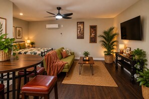 A living room with a sofa, a dining table, a coffee table, and a TV mounted on the wall. - Cozy Starkville Cottage — 4 Peaceful Acres + Pet Friendly (Starkville)