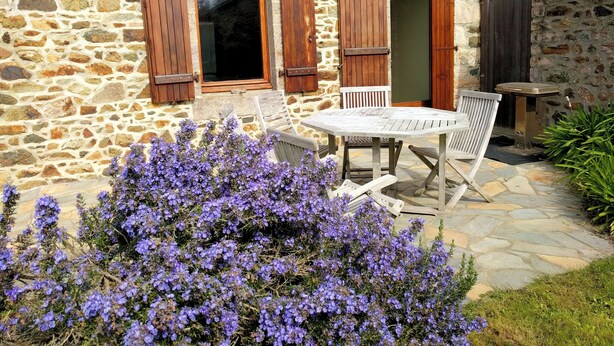 Ein Steinhaus mit Holzläden, einem runden Tisch und Stühlen, umgeben von violetten Blumen.