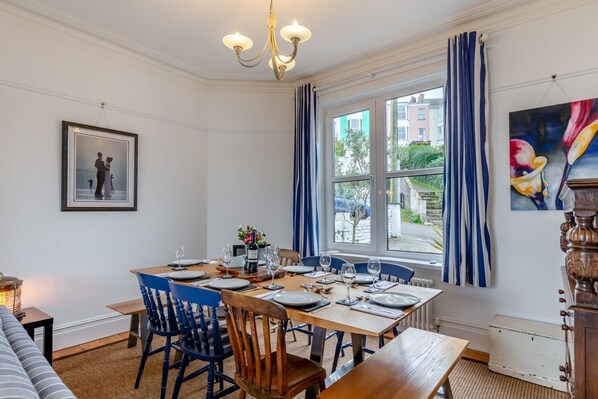 A dining room with a wooden table set for four, blue chairs, a chandelier, a large window with curtains, and a painting on the wall.