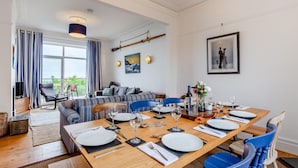 Un comedor con una mesa de madera dispuesta para seis personas, rodeada de sillas azules. Se observa un sofá y una televisión al fondo.