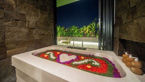 A bathtub with a flower arrangement, a window with a view of greenery, and a stone wall. - 3Bedroom-703-Private Pool Villa Sanur (Denpasar)