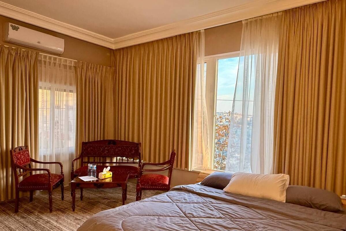 Ein Hotelzimmer mit einem großen Bett, zwei Sesseln, einem kleinen Tisch mit einer Flasche und einer Taschentuchbox sowie einem Fenster mit Vorhängen.
