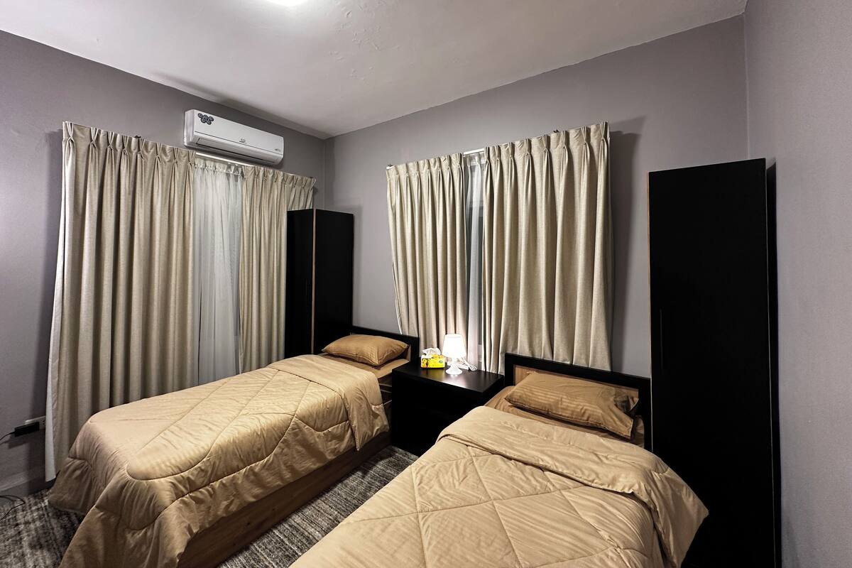 Ein Hotelzimmer mit zwei Einzelbetten, einem Nachttisch mit Lampe, einer Klimaanlage und Vorhängen.