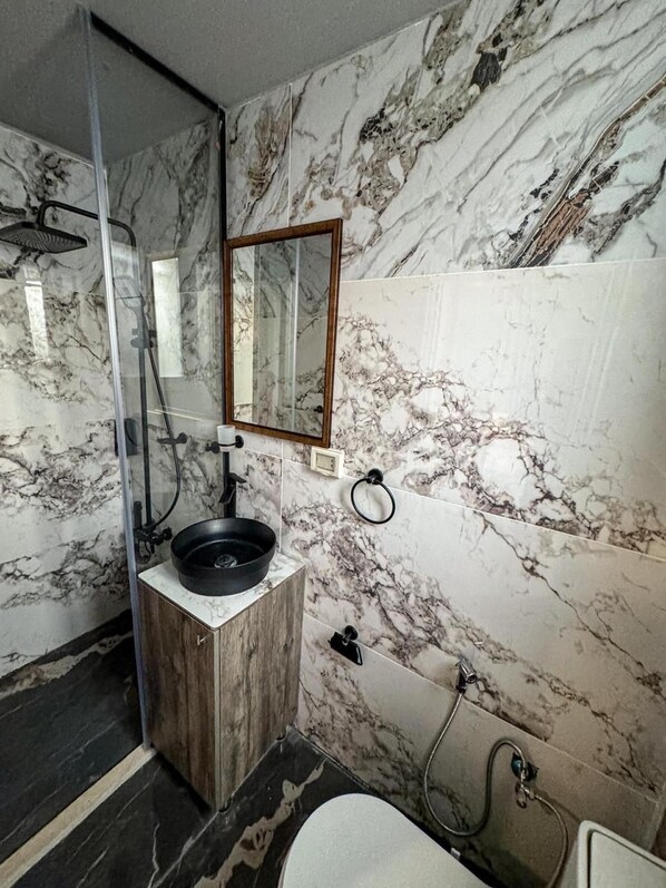 A bathroom with a marble wall, a wooden vanity, a black basin, a glass shower enclosure, and a mirror.