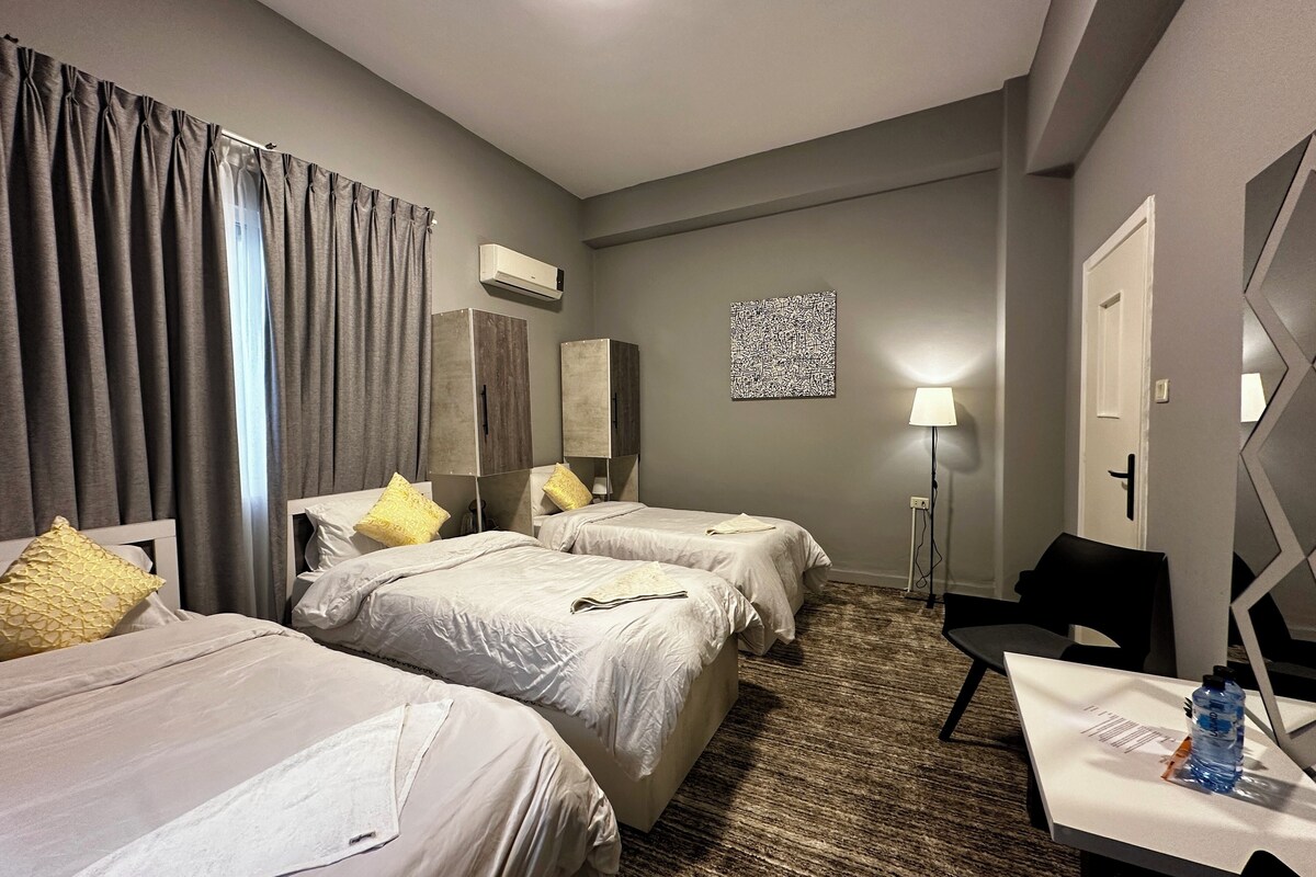 A hotel room with two beds, a desk, a chair, an air conditioning unit, a lamp, and a patterned rug.
