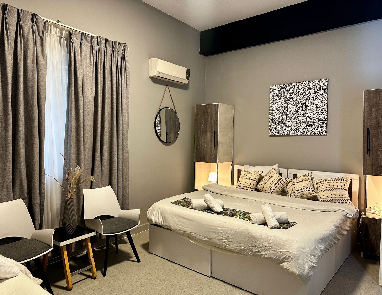 A modern bedroom with a bed, bedside tables, a chair, an air conditioning unit, and a decorative wall piece.