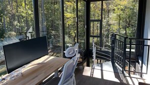A modern room with a large TV on a wooden table, a white chair, and a balcony with a glass railing overlooking a forested area. - Hacker House - Do Your Best Work (LaGrange)