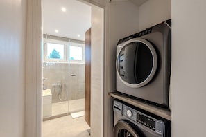 A modern laundry room with a front-loading washer and dryer, a glass shower enclosure, and a view of the outside through a window. - À Quelques Pas du Lac Léman • 2BR • Lake View • Walk to Lake & Town (Évian-les-Bains)