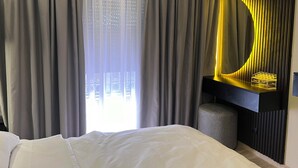 Une chambre d’hôtel comprenant un lit, une grande fenêtre avec des rideaux, un appareil de climatisation et une coiffeuse fixée au mur avec un miroir.