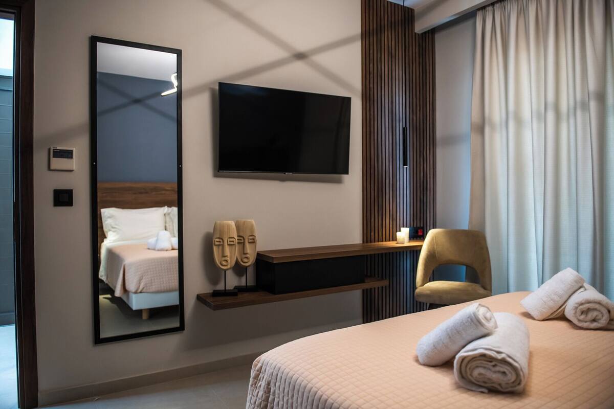 Une chambre d’hôtel avec un lit, une télévision, une chaise, un miroir et une fenêtre avec des rideaux.