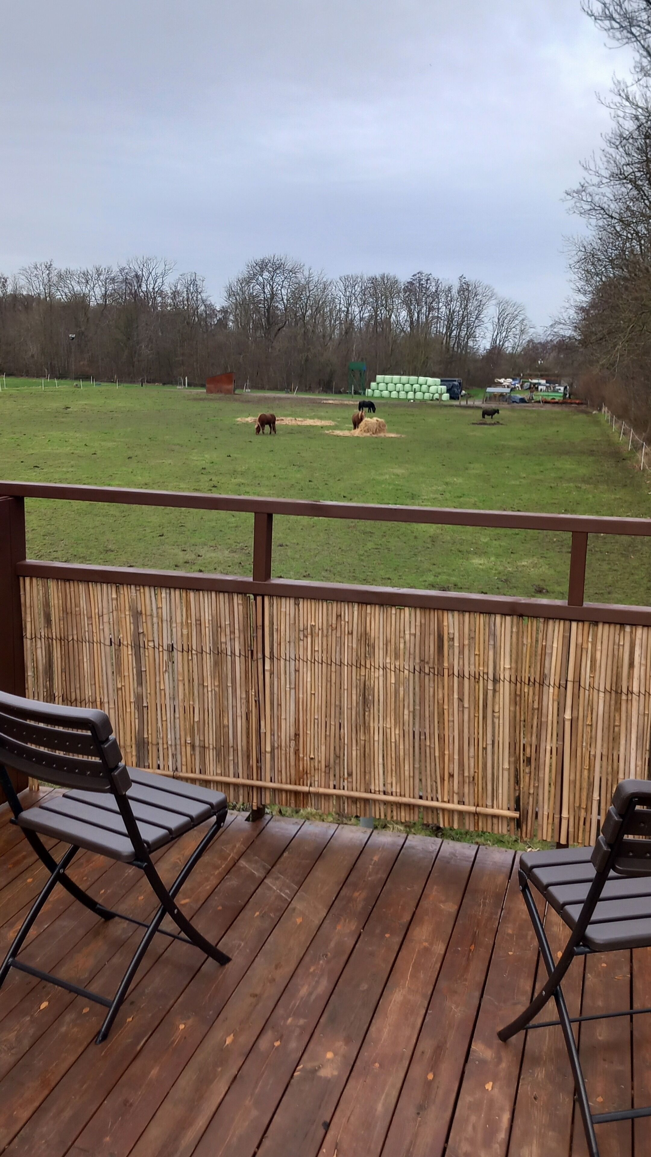 A wooden deck with two black chairs, overlooking a grassy field with horses and hay bales.