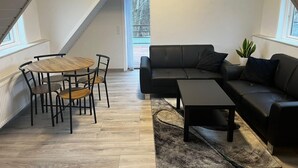 A loft-style room with a wooden dining table and chairs, a black leather sofa, and a small coffee table. There is a window with a view of trees outside. - Sønderborg in Denmark (Sønderborg)