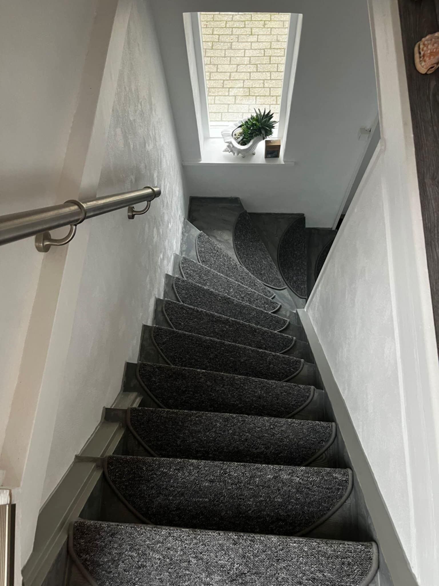 Un escalier avec une rampe, des marches recouvertes de moquette et une petite fenêtre ornée d’une plante.