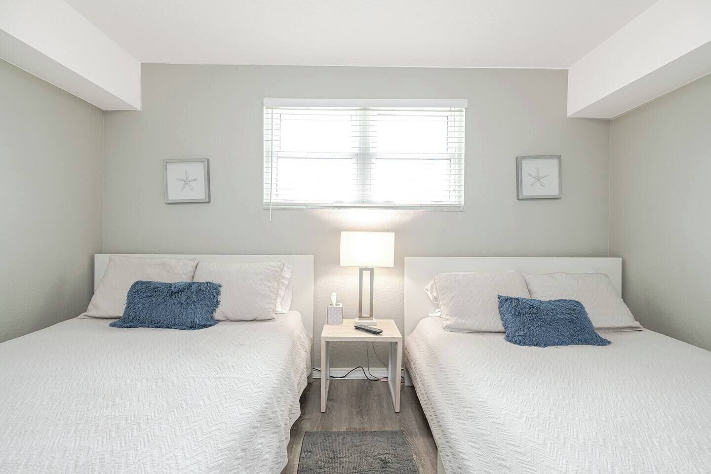 A twin bedroom with two beds, a bedside table with a lamp, and a window with blinds.