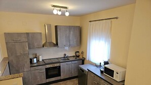 Een kleine keuken met grijze kastjes, een kookplaat, oven, magnetron en een raam met gordijnen.