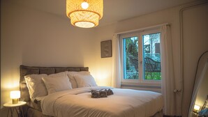 Un dormitorio con una cama grande, una lámpara de noche, una ventana con cortinas y vistas al exterior con vegetación.