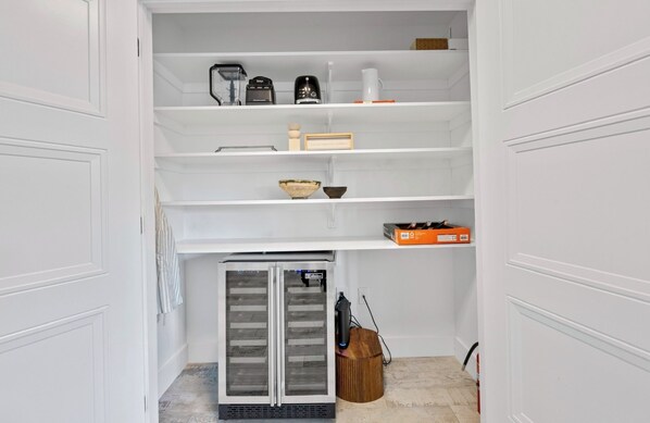 A white interior with a built-in refrigerator, shelves with kitchen appliances, and a wooden basket.