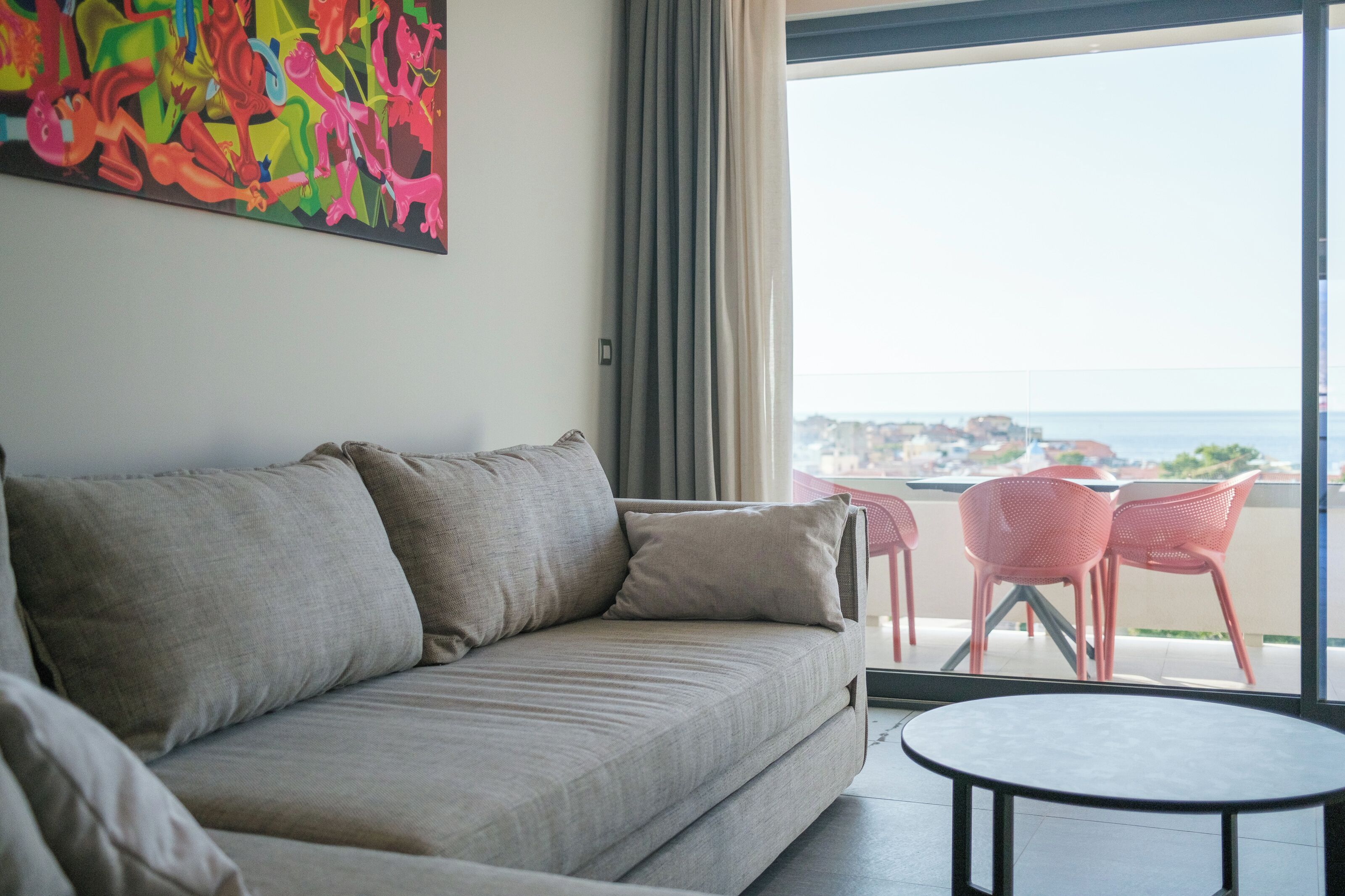 A modern living room with a beige sofa, a colorful abstract painting, and a view of the sea through large windows.