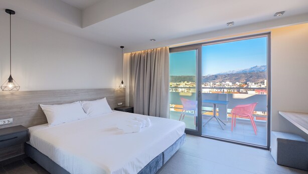 A modern hotel room with a large bed, a nightstand, a desk, and a balcony with a view of the city and mountains. - Sky-High 70sqm Penthouse w. Stunning Views - 05

 (Chania)