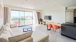 A modern living room with a sofa, a dining table with chairs, a TV, and a kitchen area with a refrigerator. - Sky-High 70sqm Penthouse w. Stunning Views - 05

 (Chania)