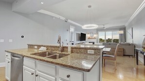 A modern kitchen with granite countertops, a stainless steel sink, and white cabinetry. There is a dining area with a table and chairs, and a large window offering a view of the outdoors.