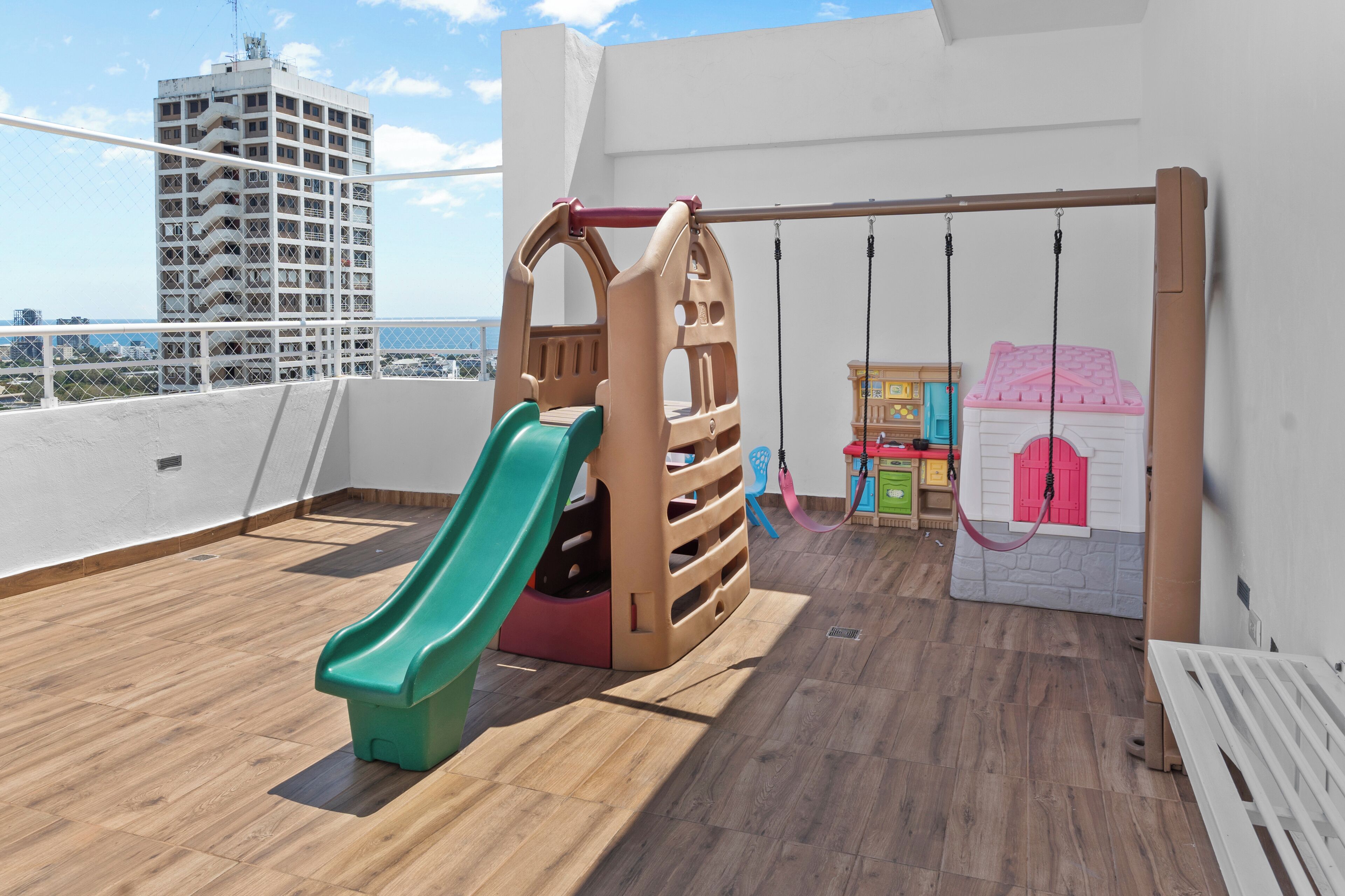 A children's play area with a slide, swings, and a playhouse on a balcony with wooden flooring and a view of a tall building.
