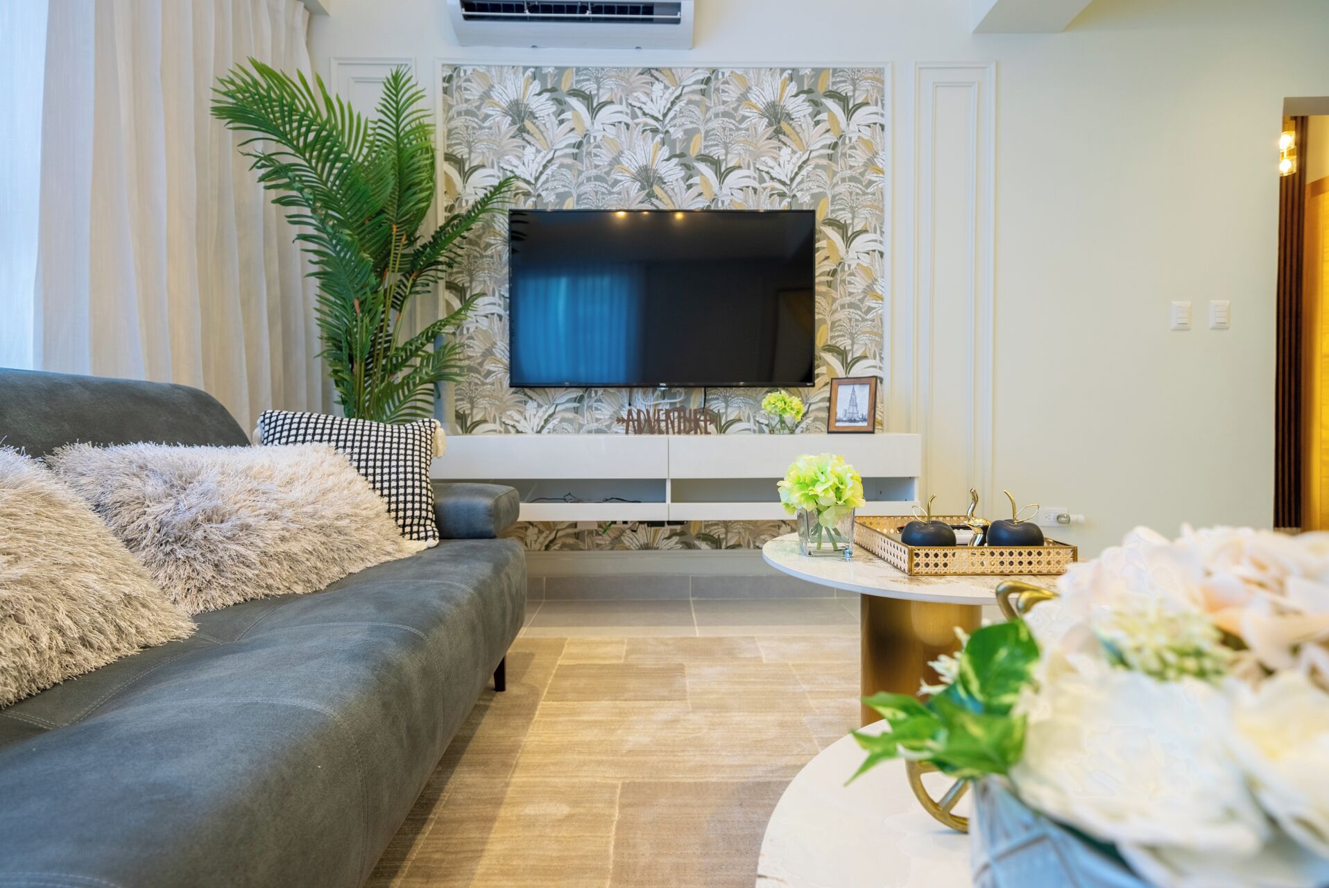 A living room with a grey sofa, a flat-screen TV mounted on the wall, a large potted plant, and a small round table with decorative items.