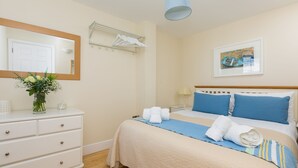 Un letto rifatto con precisione, lenzuola nei toni del blu e del beige, una cassettiera bianca con cassetti, uno specchio, un quadro incorniciato appeso al muro e un appendiabiti.