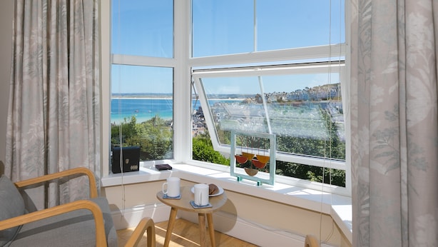 Una stanza con una grande finestra che si affaccia sul mare e sugli edifici. Si vede una sedia di legno, un tavolino con tazza e piattino, e un quadro incorniciato sul davanzale.