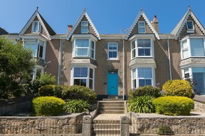 A row of three-story houses with bay windows, a central blue door, and a stone wall with a metal gate. - Captain’s Rest, St Ives – Sleeps 4 – Panoramic Views Dog-Friendly – Parking (St Ives)
