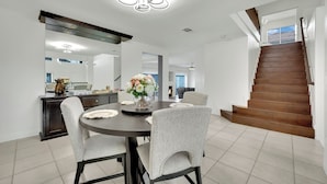 Une cuisine moderne avec un plan de travail foncé, des armoires blanches et une table de salle à manger ronde entourée de quatre chaises. Un lustre est suspendu au-dessus de la table, et un vase rempli de fleurs est posé sur la table. Un escalier en bois mène à l’étage supérieur.