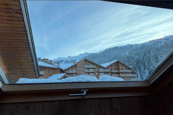 Vista a través de un tragaluz de un edificio de estilo chalé con varios balcones, rodeado de árboles y montañas nevadas.