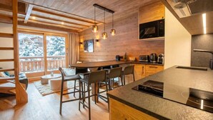 A modern kitchen with wooden accents, a bar area, and a view of snow-covered trees. - Troillet 11, 200m away from the ski pistes (Les Allues)